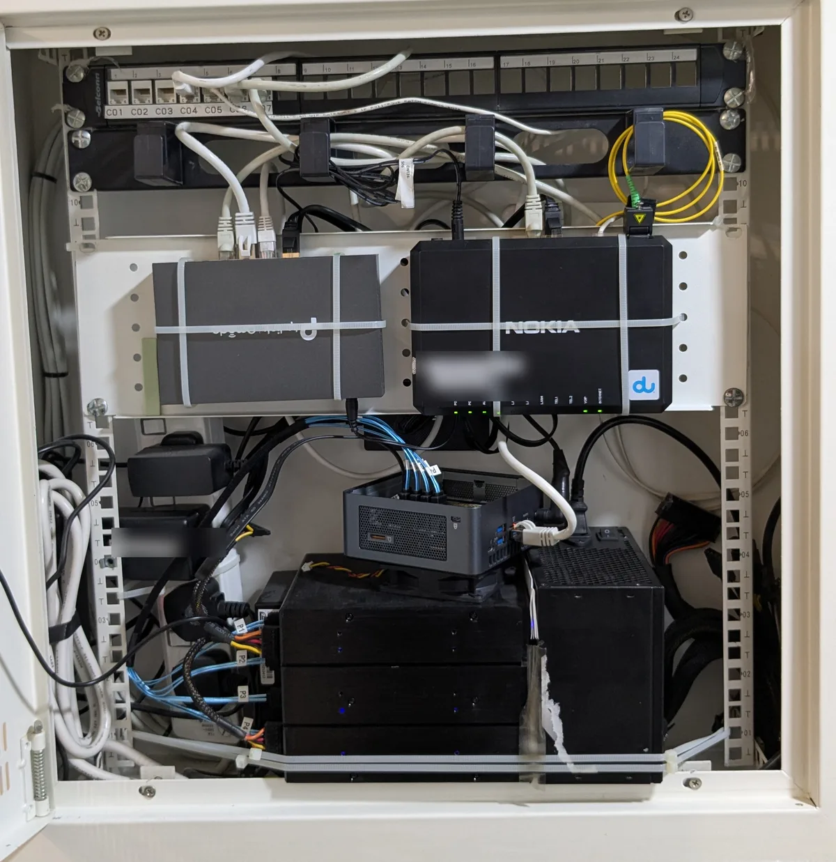The wall rack: patch panel on top, router and fiber ONT on the shelf, and under it the NUC sitting on a 4-bay drive enclosure with the ATX PSU beside it and blue SATA cables running from the NUC to the drives